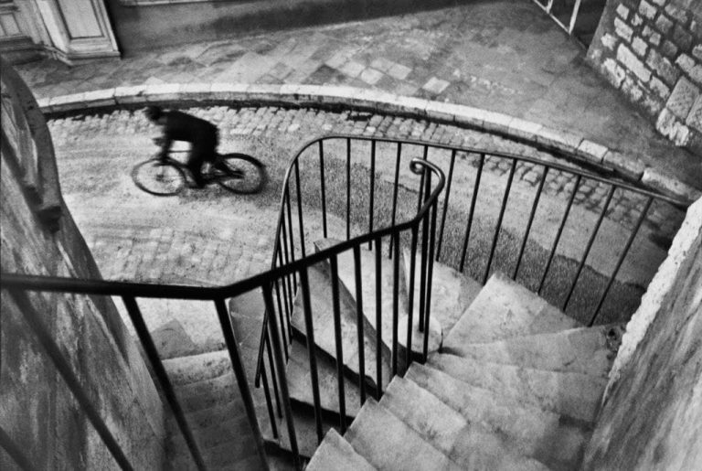 Fotografía de Henri Cartier-Bresson en Hyères, Francia, 1932: ciclista pasando bajo una escalera en espiral, composición geométrica